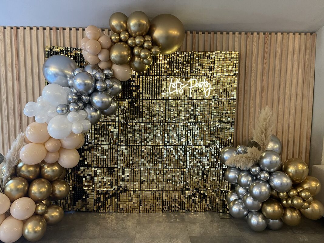 Backdrop à sequins or et néon « Let’s Party » devant un mur à lattes de bois, encadrés de grappes de ballons or, argent, blancs et nude et de pampas