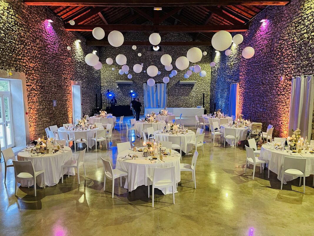 Salle aux murs de pierre sous charpente : dizaines de lampions blancs suspendus, murs éclairés d’orange et de violet, tables rondes nappées de blanc