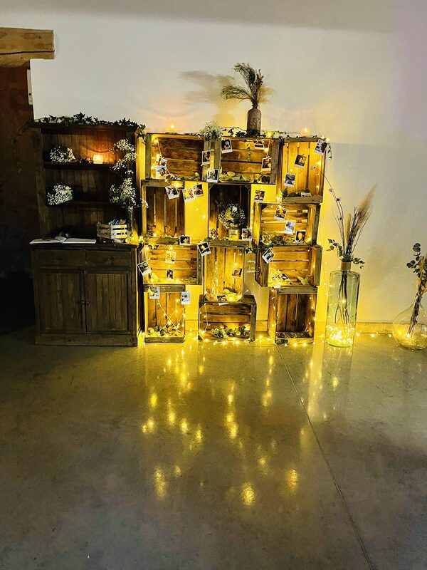 Mur de caisses en bois empilées traversé de guirlandes LED et de photos, buffet en bois garni de gypsophile, dames-jeannes aux pampas