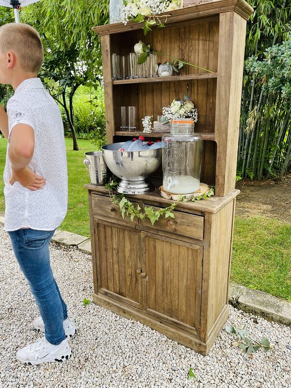 Buffet en bois dans le jardin pour le vin d’honneur : fontaine à boisson sur rondin, verres, gypsophile et roses blanches, guirlande de lierre
