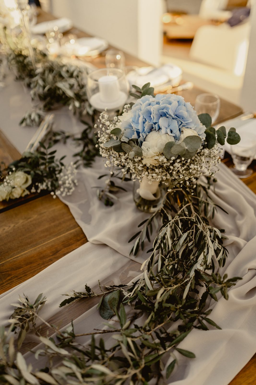Bouquet d’hortensias bleus et gypsophile sur un chemin de voile gris, branches d’olivier et bougie sur table en bois