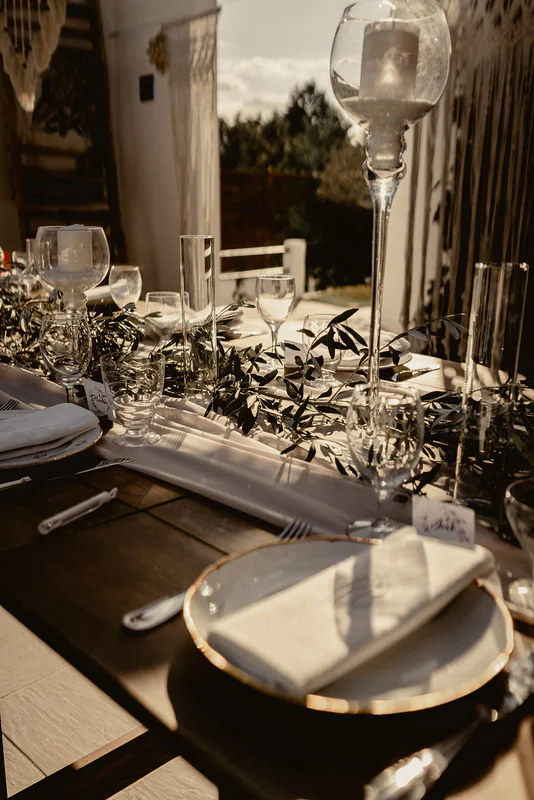 Table dressée au soleil couchant : assiettes à liseré doré, serviettes blanches, olivier et bougies sur hauts pieds de verre