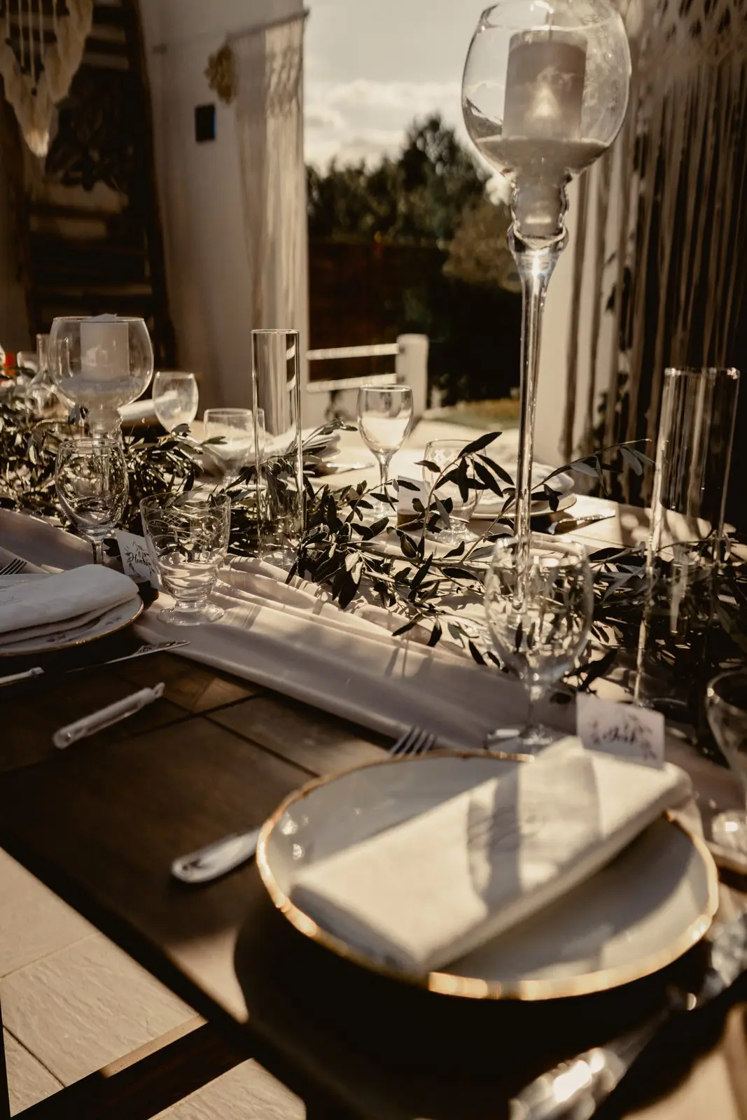 Table dressée au soleil couchant : assiettes à liseré doré, serviettes blanches, olivier et bougies sur hauts pieds de verre