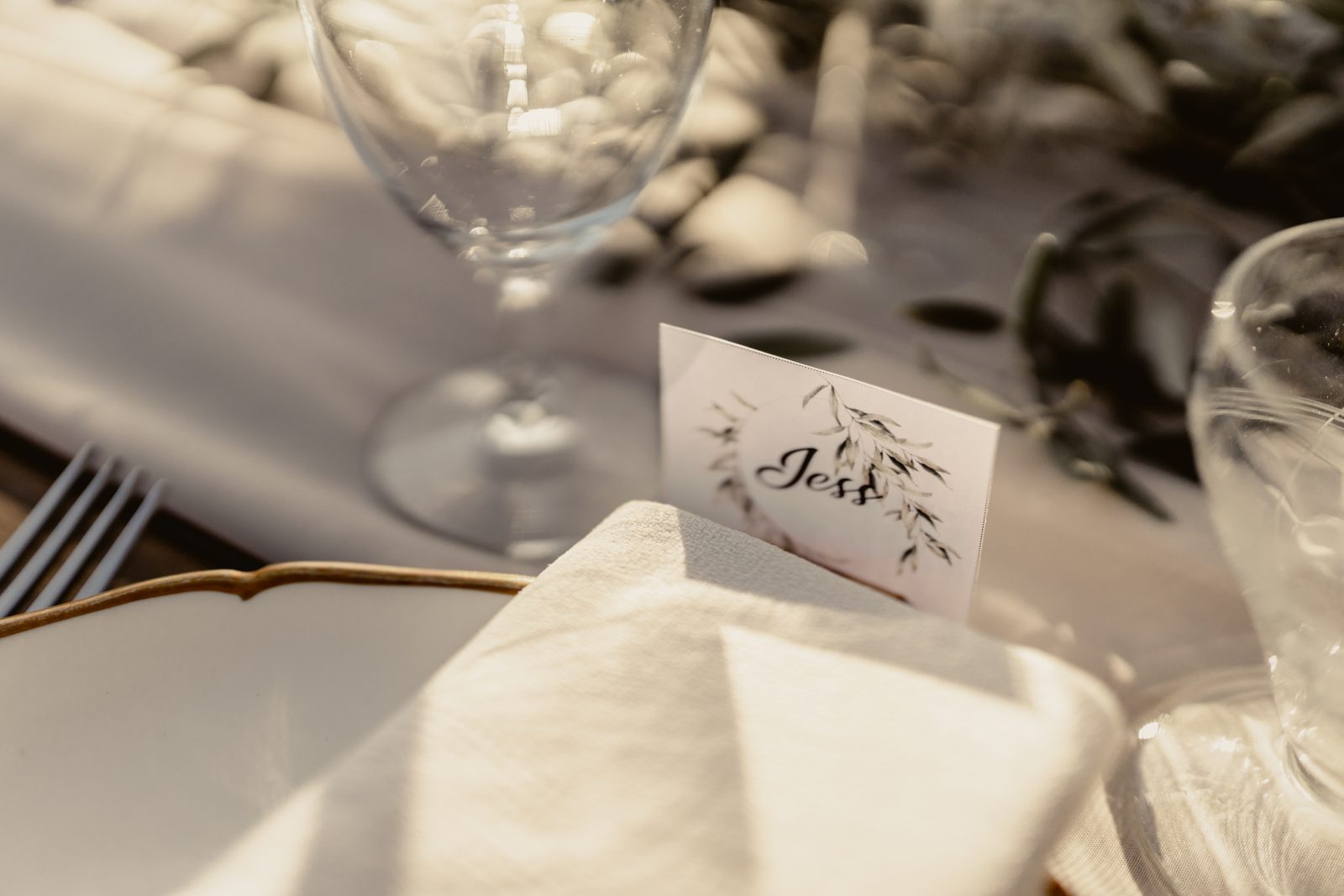 Gros plan d’un marque-place illustré de feuillage posé sur une serviette blanche, assiette à liseré doré et verres