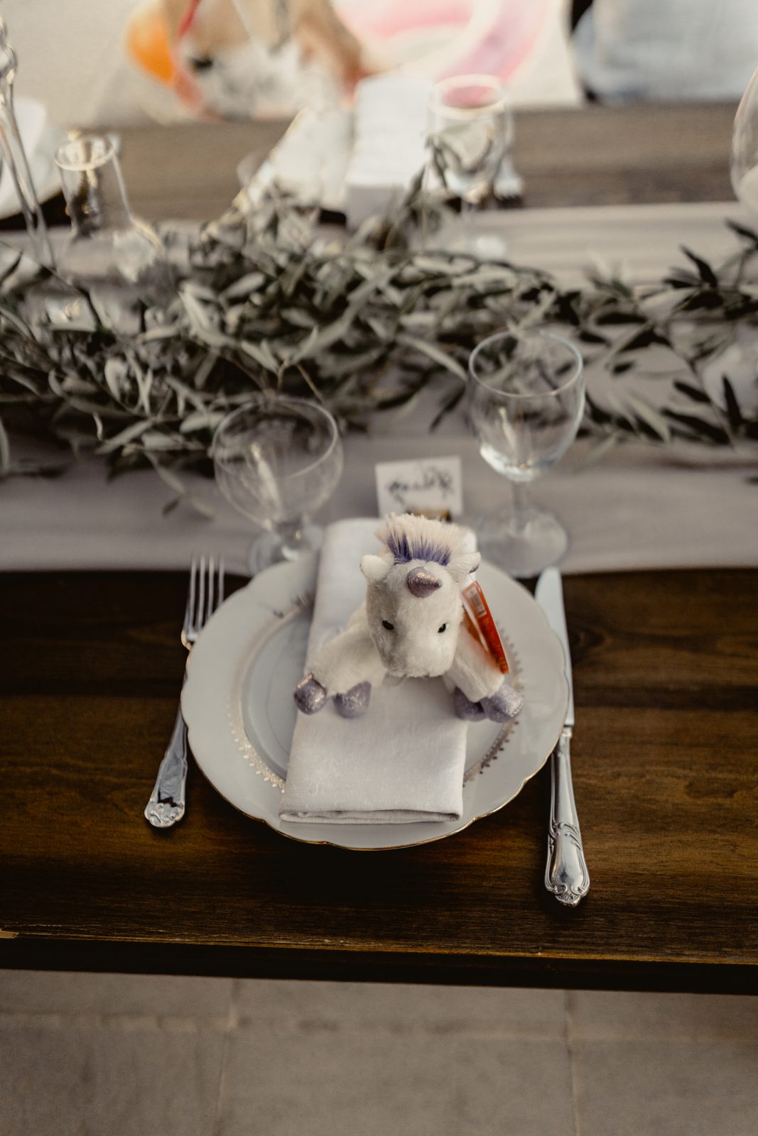 Couvert d’enfant sur table en bois : assiette blanche, serviette en coton, peluche licorne et branches d’olivier