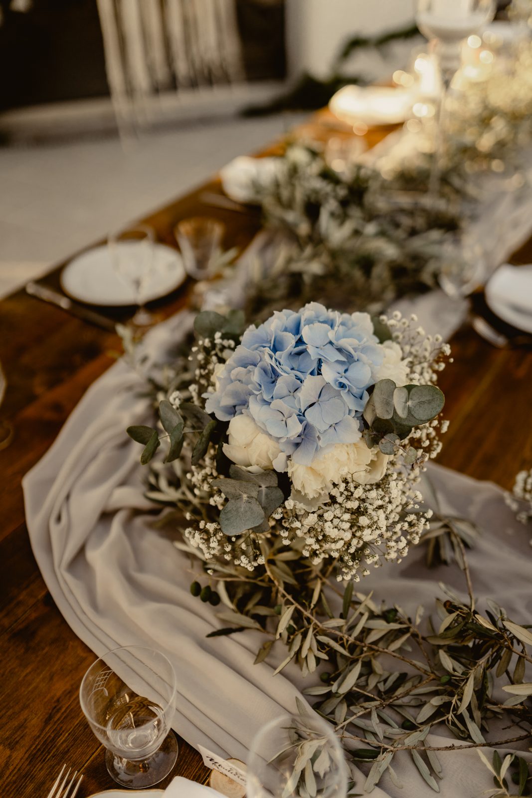 Centre de table d’hortensias bleus, roses blanches et gypsophile sur voile gris et olivier, table en bois au crépuscule