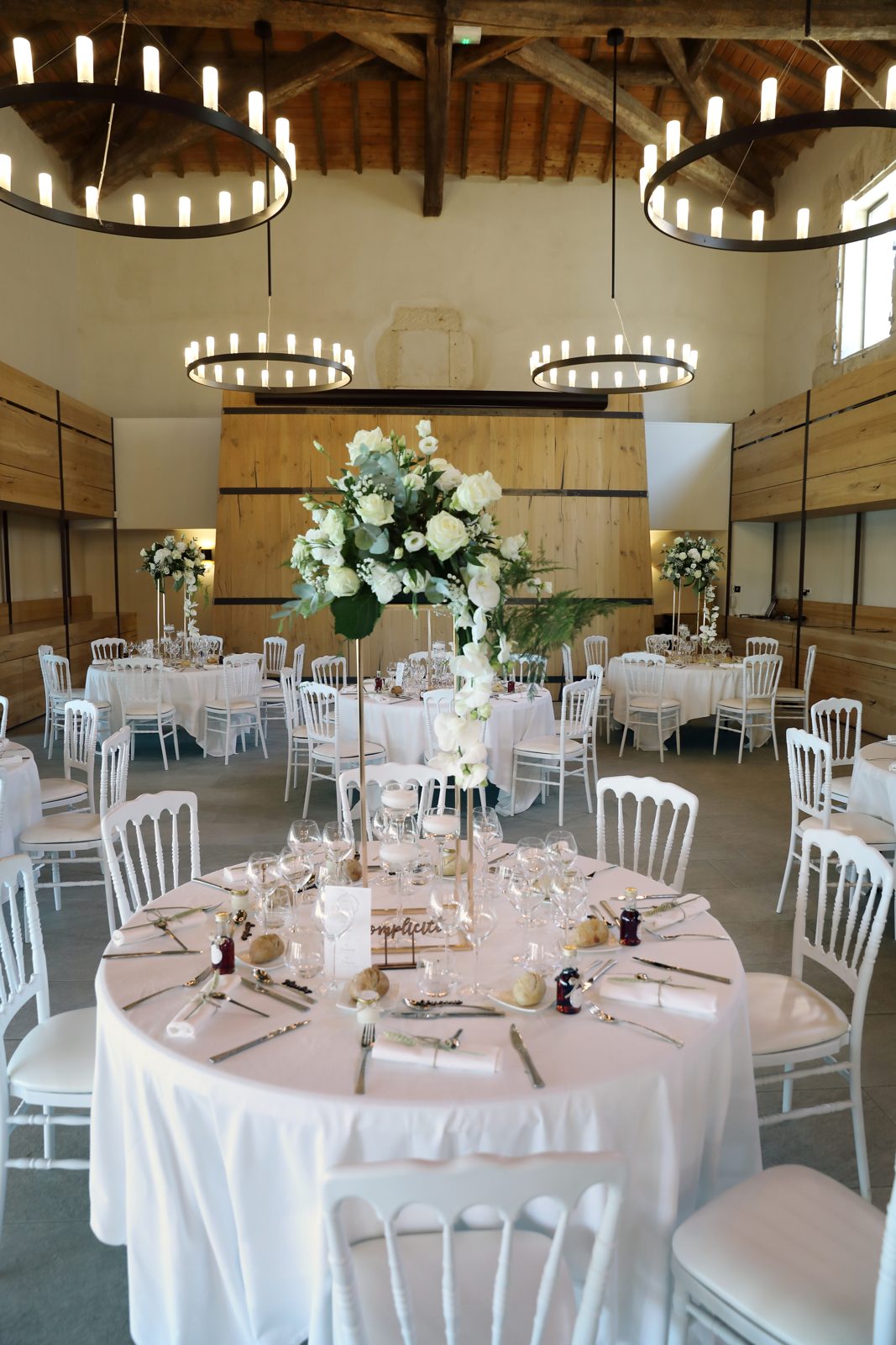 Salle aux lustres à bougies, table ronde nappée de blanc, chaises Napoléon III blanches et haut bouquet de roses blanches