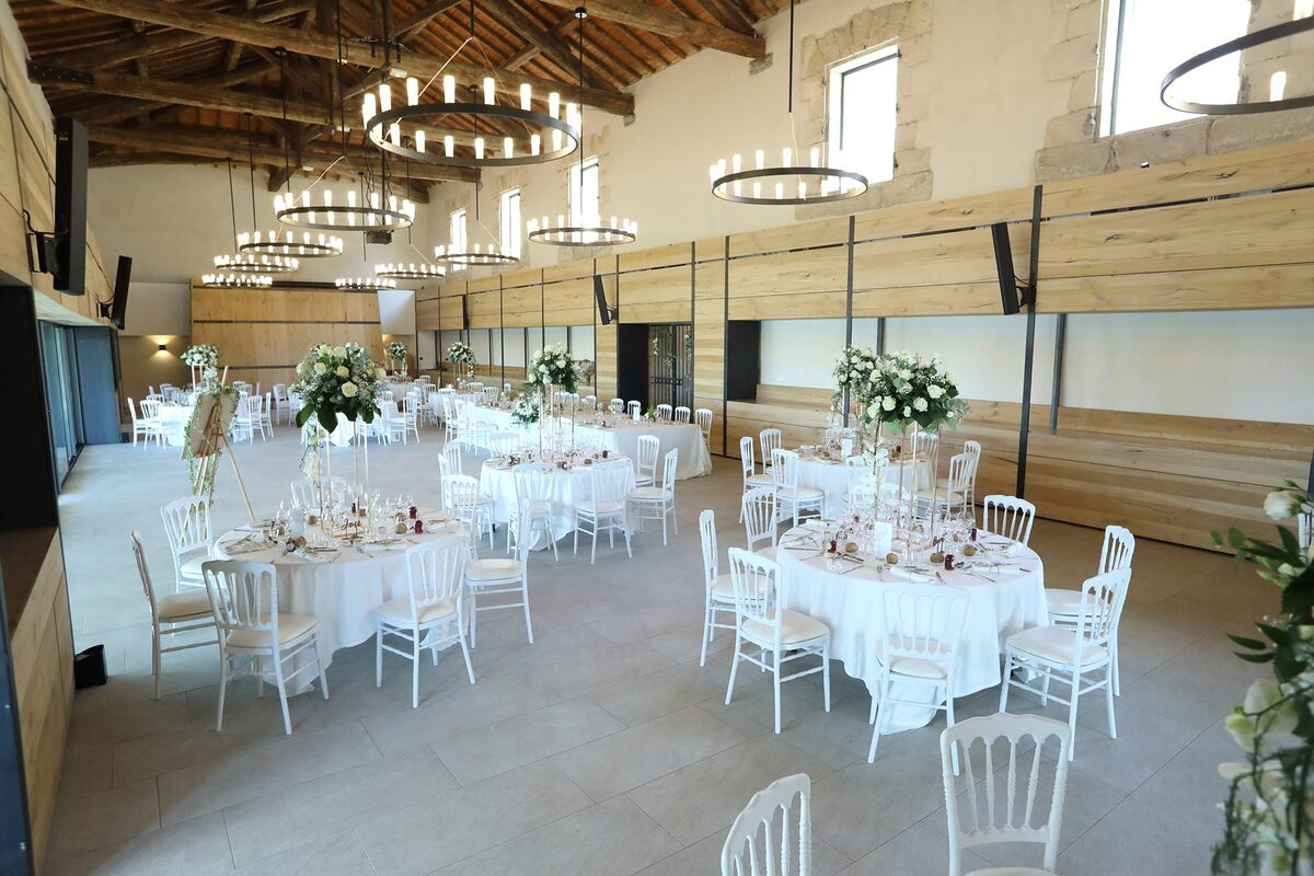 Grande salle aux lustres circulaires, tables rondes nappées de blanc, chaises Napoléon III blanches et hauts bouquets blancs