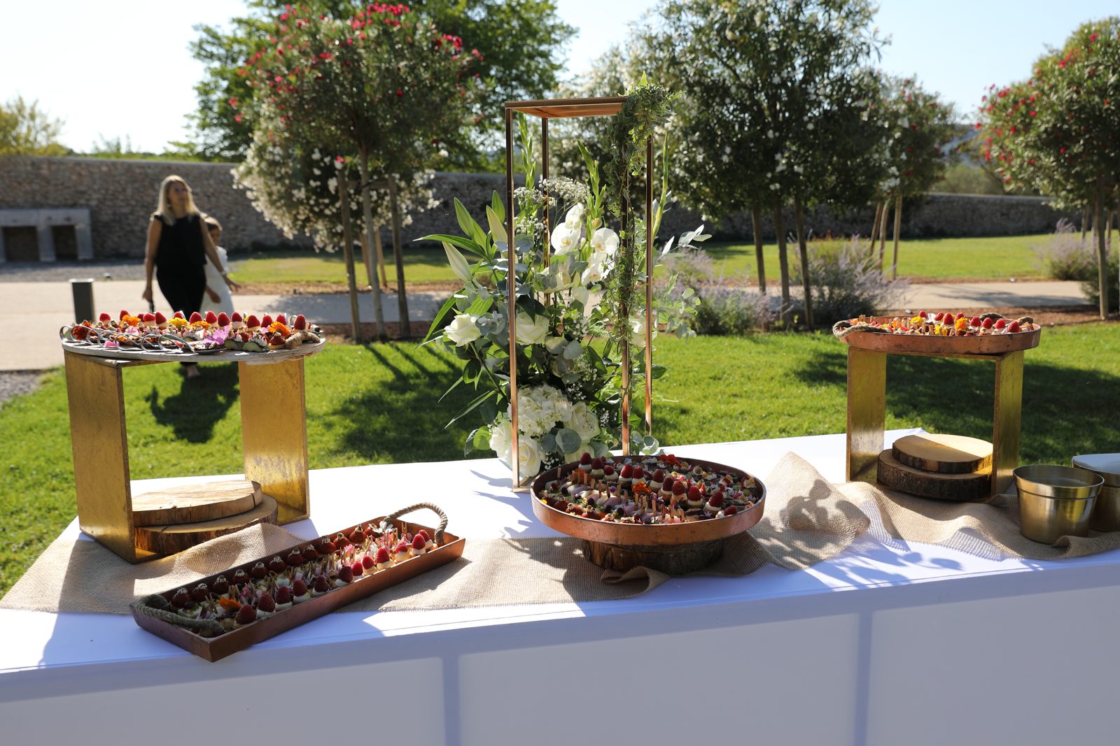 Buffet de desserts en extérieur sur supports dorés et plateaux en bois, composition de fleurs blanches sur structure dorée