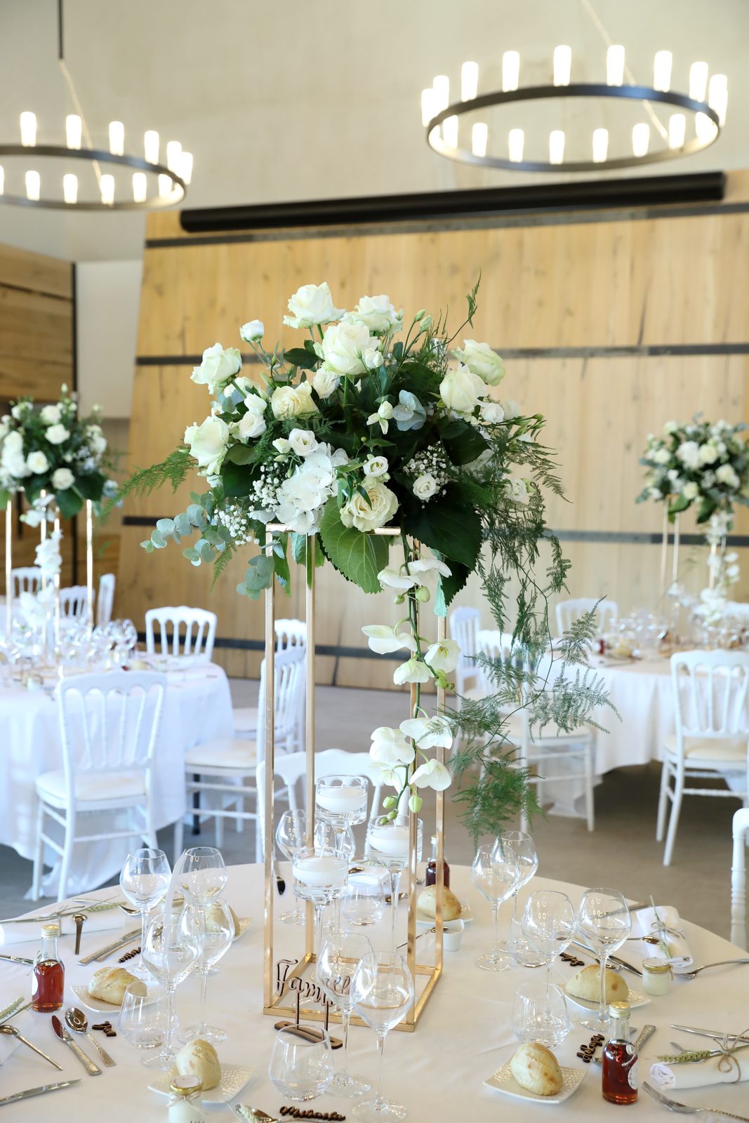 Centre de table haut de roses et orchidées blanches sur support doré, tables nappées de blanc sous des lustres à bougies