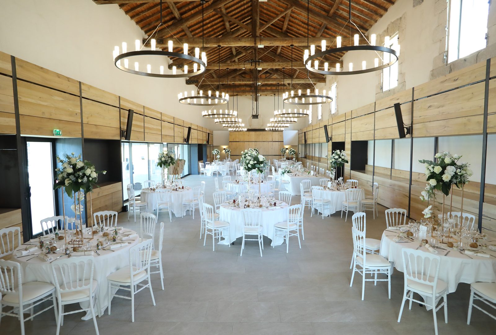 Salle aux poutres apparentes et lustres à bougies, tables rondes nappées de blanc et chaises Napoléon III blanches