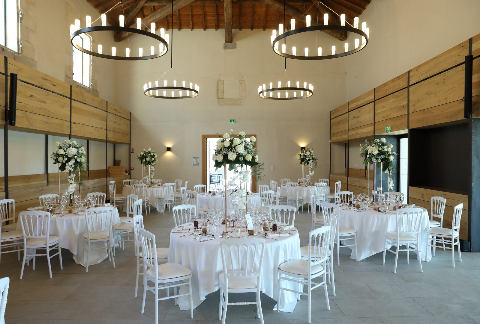 Salle de réception claire, tables rondes blanches, chaises Napoléon III blanches et hauts bouquets de roses blanches