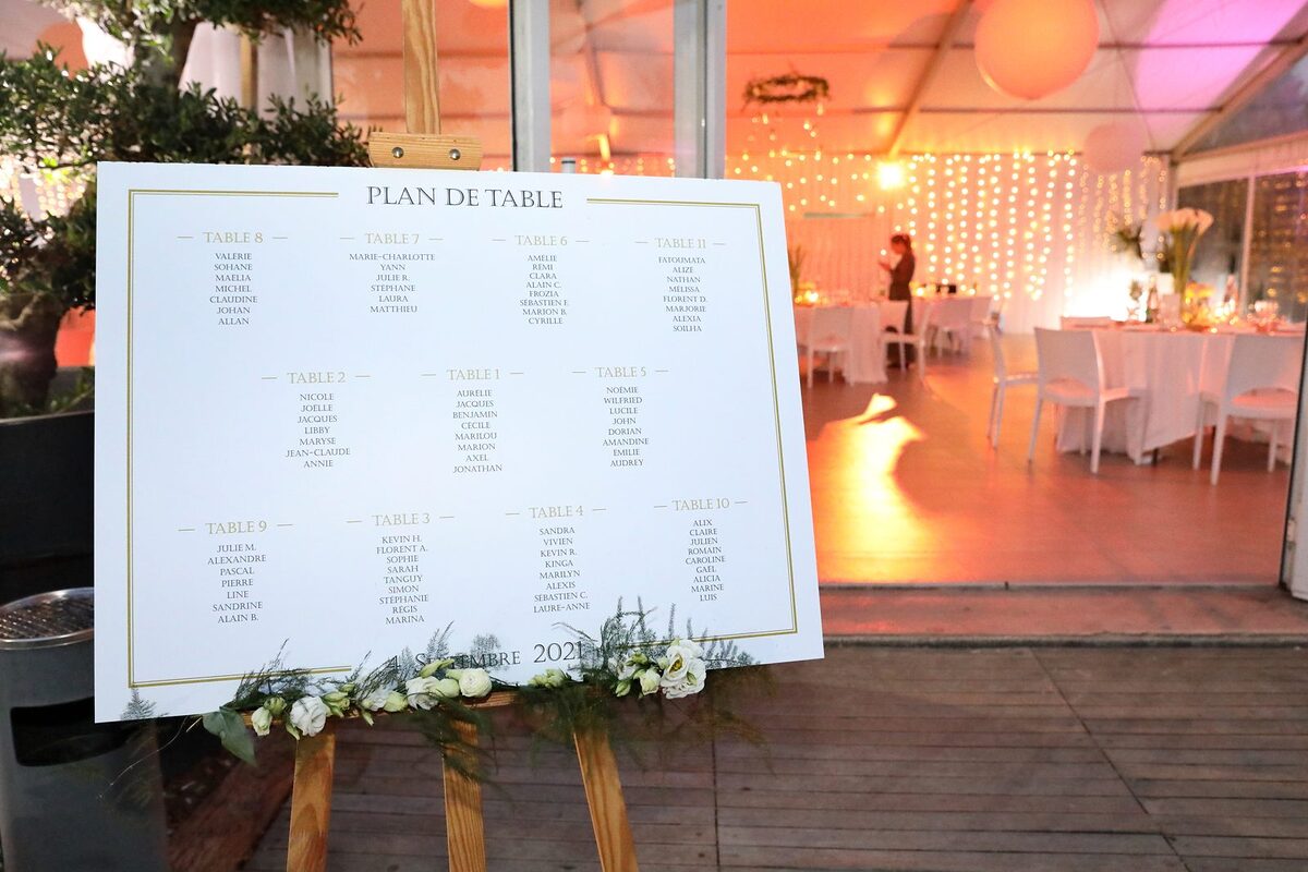Plan de table fleuri de roses blanches sur grand chevalet en bois, à l’entrée d’un chapiteau éclairé de guirlandes