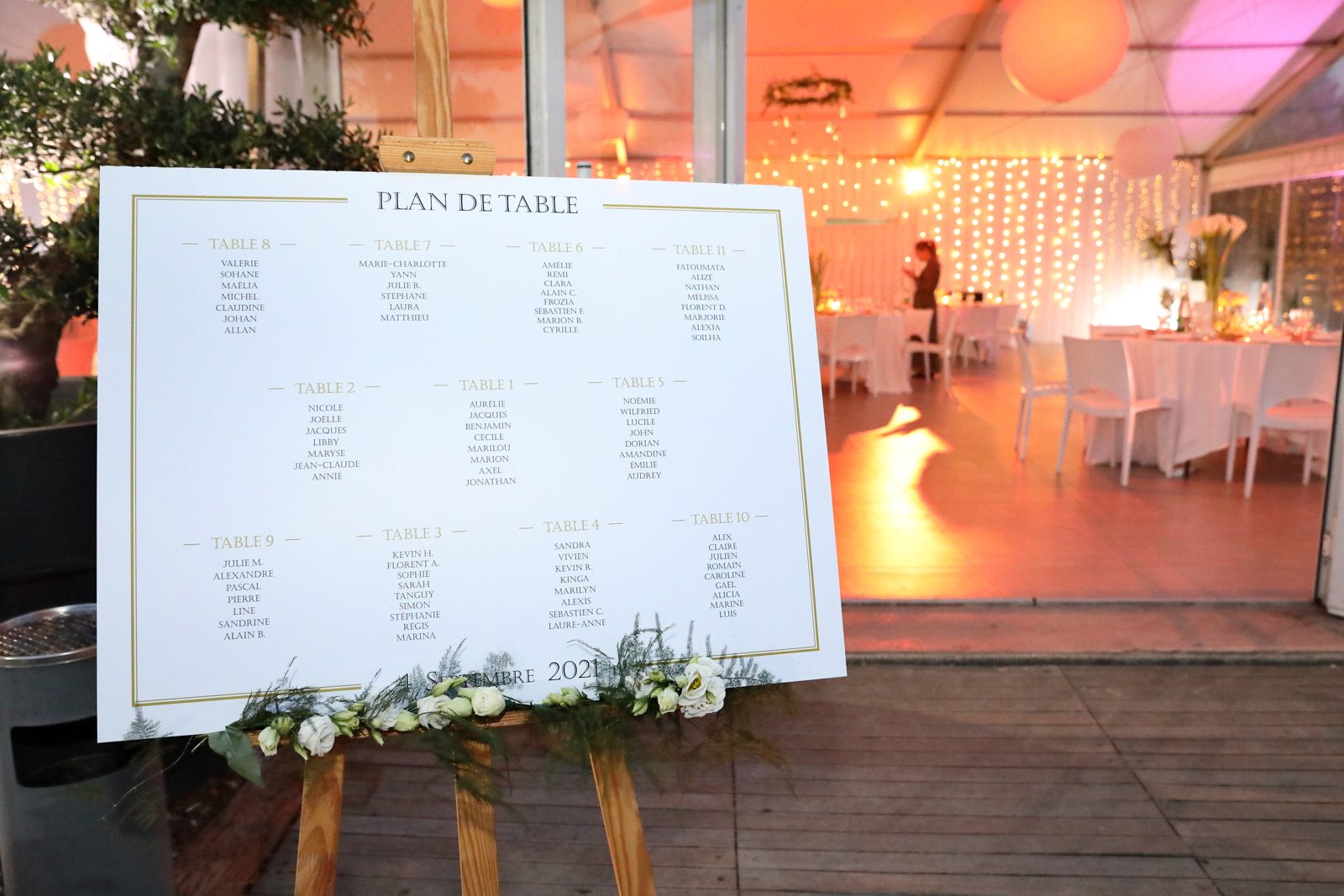 Plan de table fleuri de roses blanches sur grand chevalet en bois, à l’entrée d’un chapiteau éclairé de guirlandes