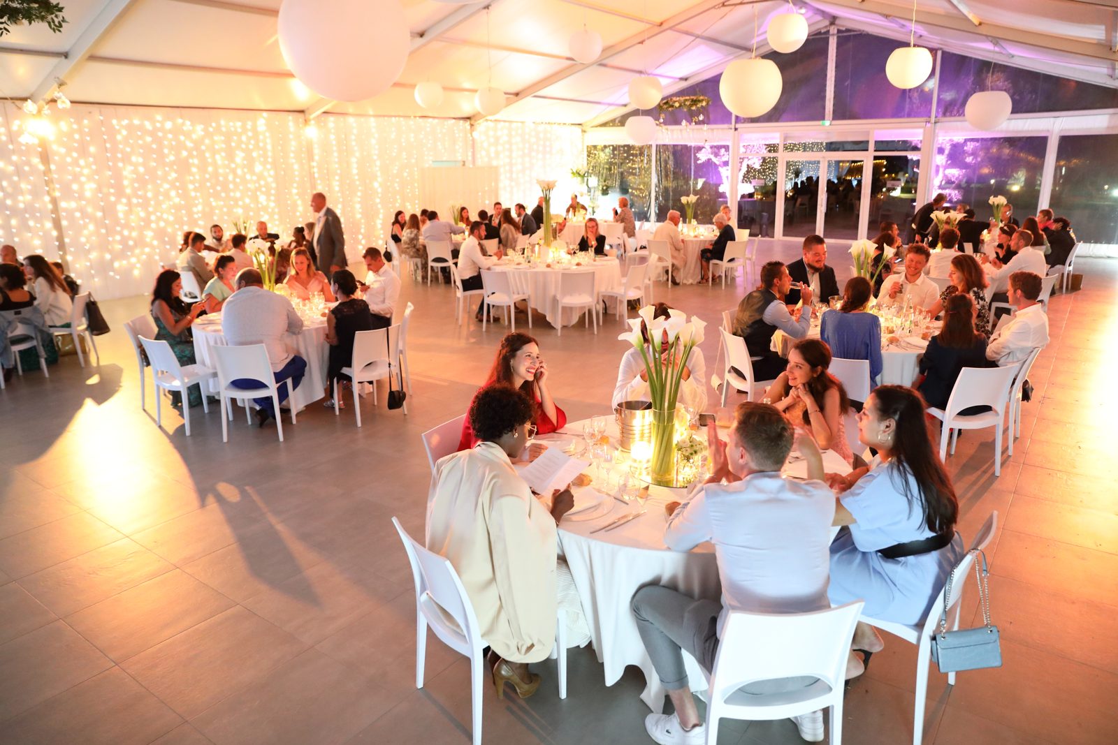 Dîner sous chapiteau éclairé, invités à des tables rondes nappées de blanc, boules lumineuses et rideaux de guirlandes