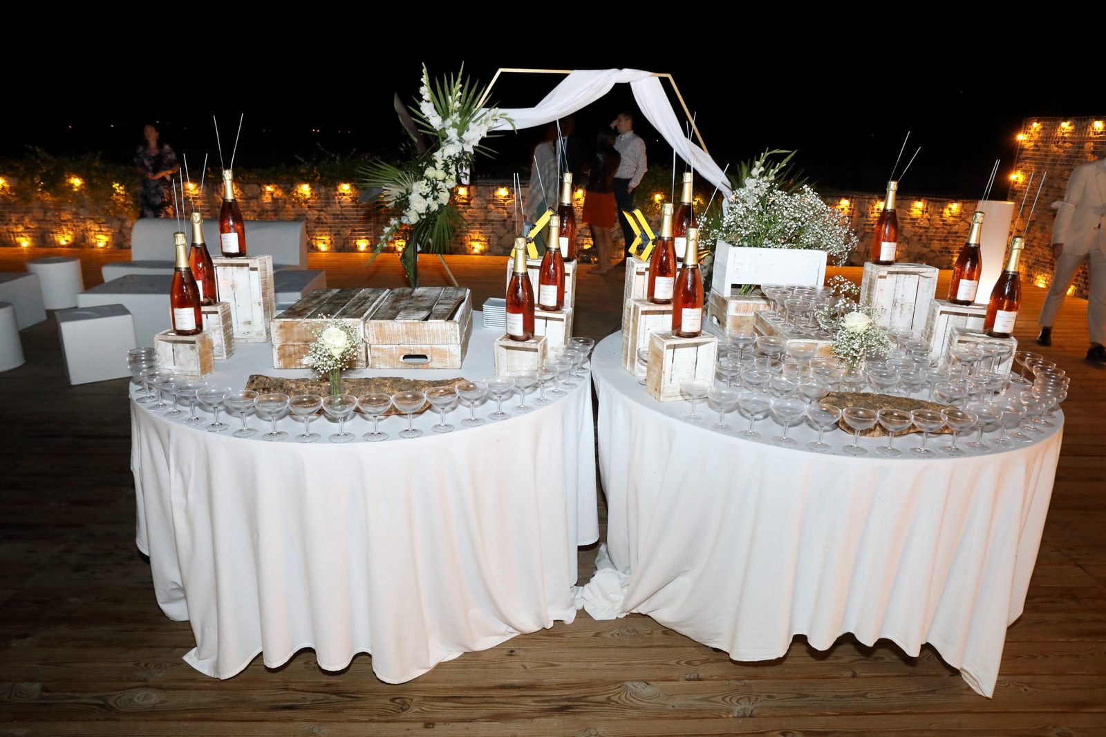Bar à champagne de nuit sur deux tables rondes nappées de blanc, caisses en bois, flûtes et bouquets blancs
