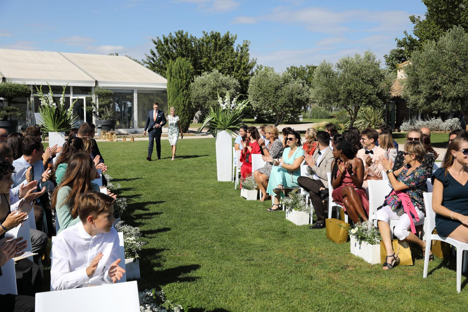Cérémonie laïque sur la pelouse, allée bordée de caisses fleuries et de hauts vases blancs, invités sur chaises blanches