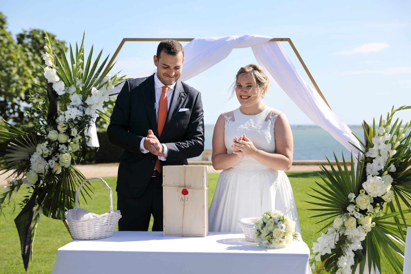 Les mariés sous une arche hexagonale dorée drapée de voile blanc, entre deux compositions de palmes et roses blanches