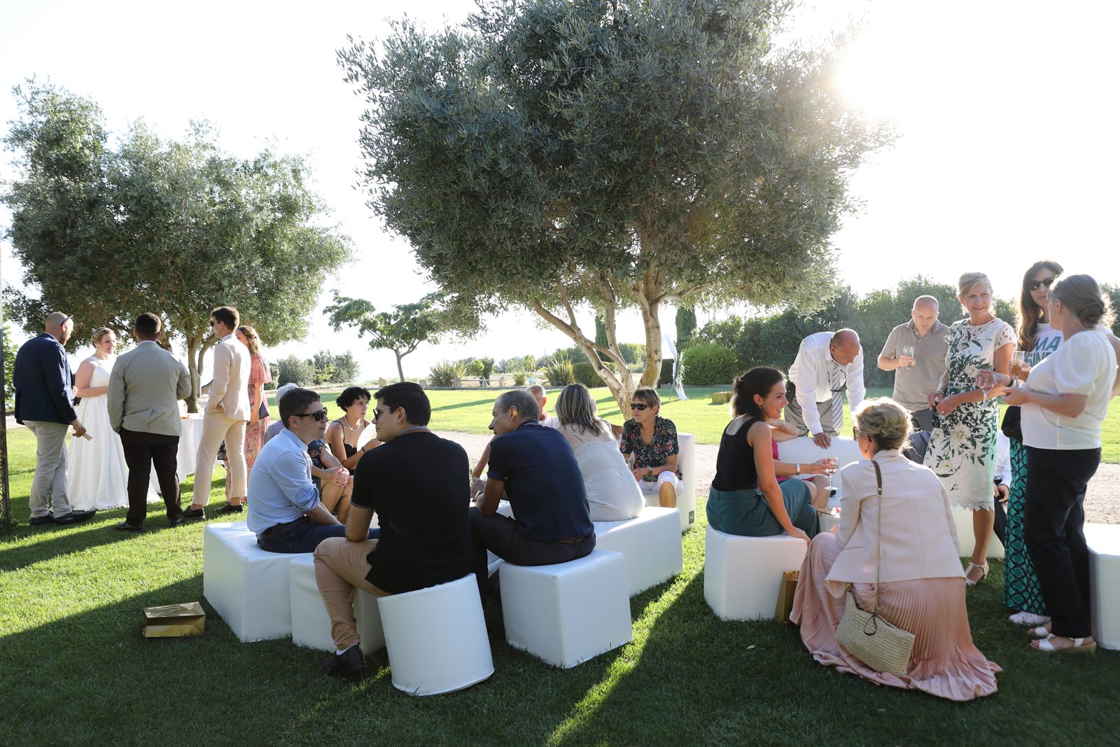 Cocktail à l’ombre des oliviers, invités assis sur des poufs cubes blancs disposés sur la pelouse