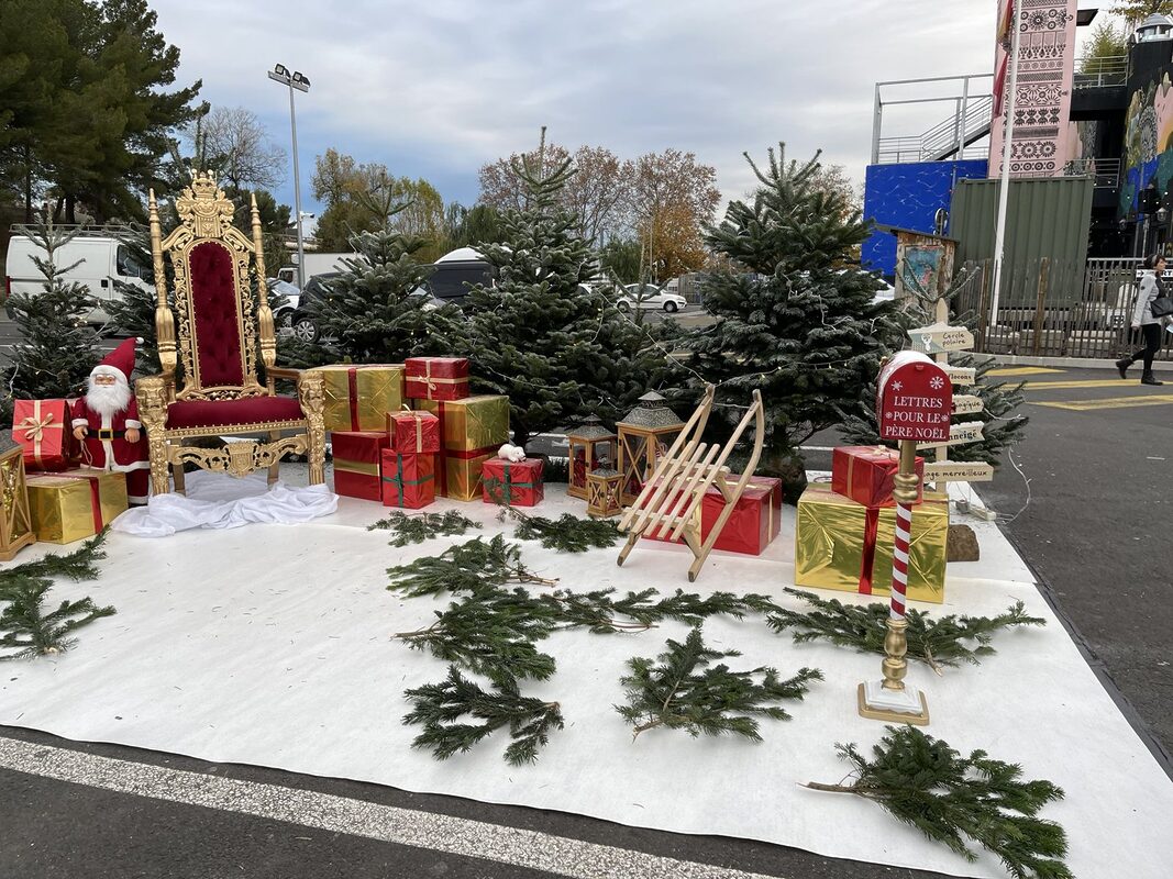 Village du Père Noël en plein air : trône doré en velours rouge, sapins naturels givrés, gros cadeaux rouges et dorés, lanternes en bois, luge et boîte aux lettres