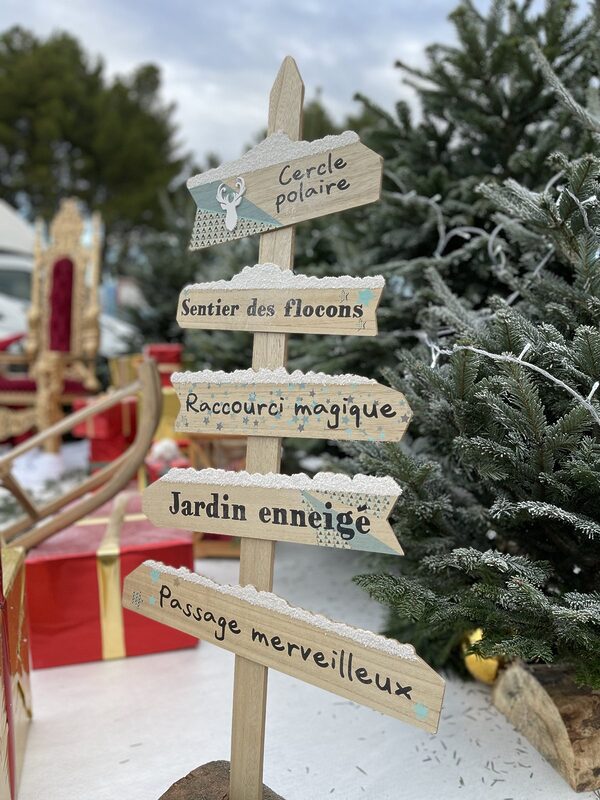 Panneau directionnel en bois « Cercle polaire, Sentier des flocons, Raccourci magique, Jardin enneigé, Passage merveilleux » près d’un sapin givré