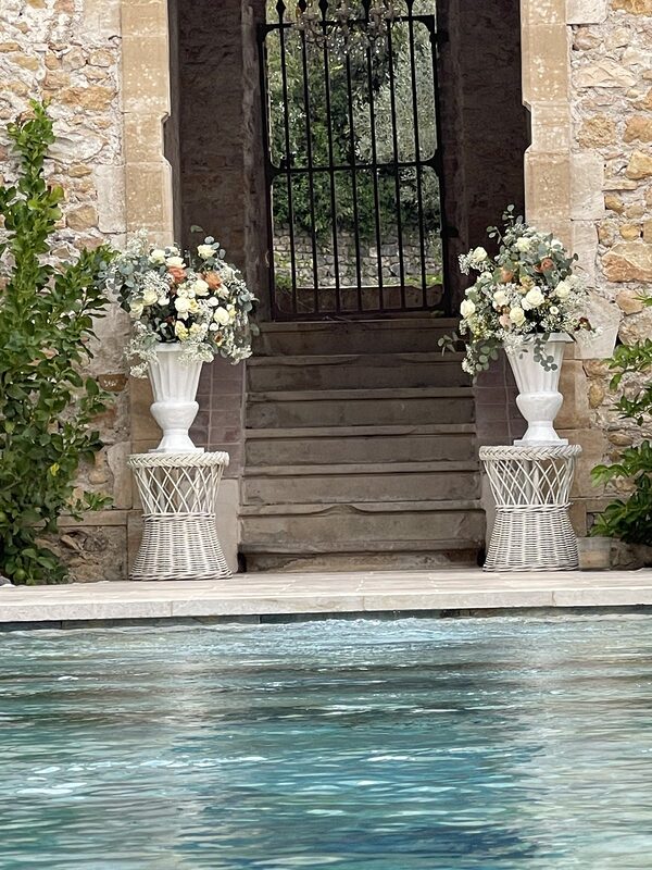 Deux grands vases Médicis blancs fleuris sur des sellettes en osier blanc, de part et d’autre d’un escalier en pierre, au bord de la piscine