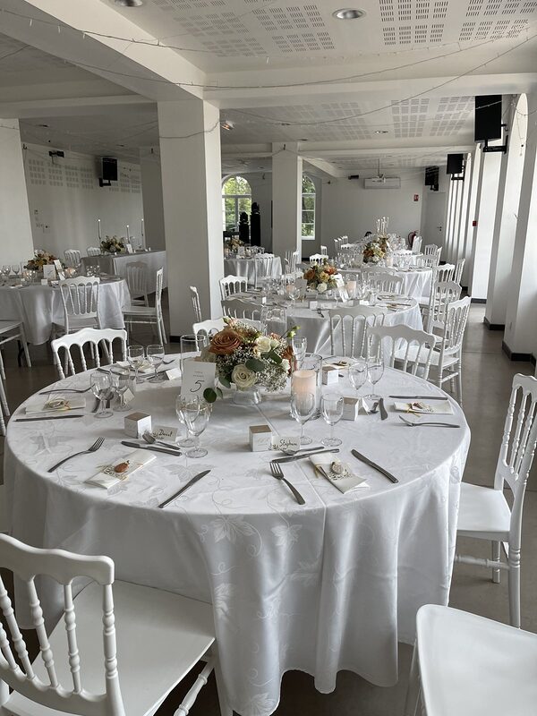 Salle de réception blanche : tables rondes nappées de blanc, chaises Napoléon III blanches, centres de table fleuris et bougies LED dans des vases cylindres