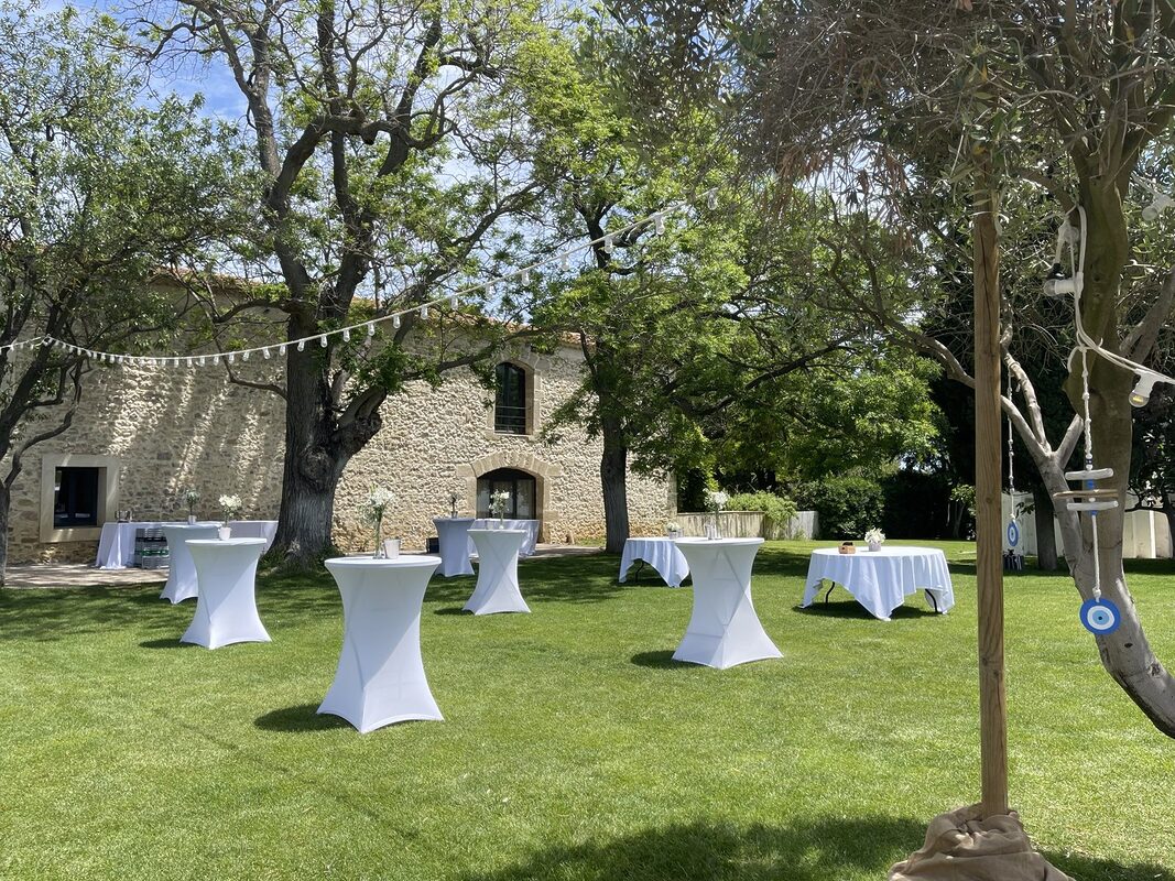 Table d’accueil nappée de blanc sous une pergola en fer, dame-jeanne d’hortensias blancs et caisse en bois fleurie à l’entrée