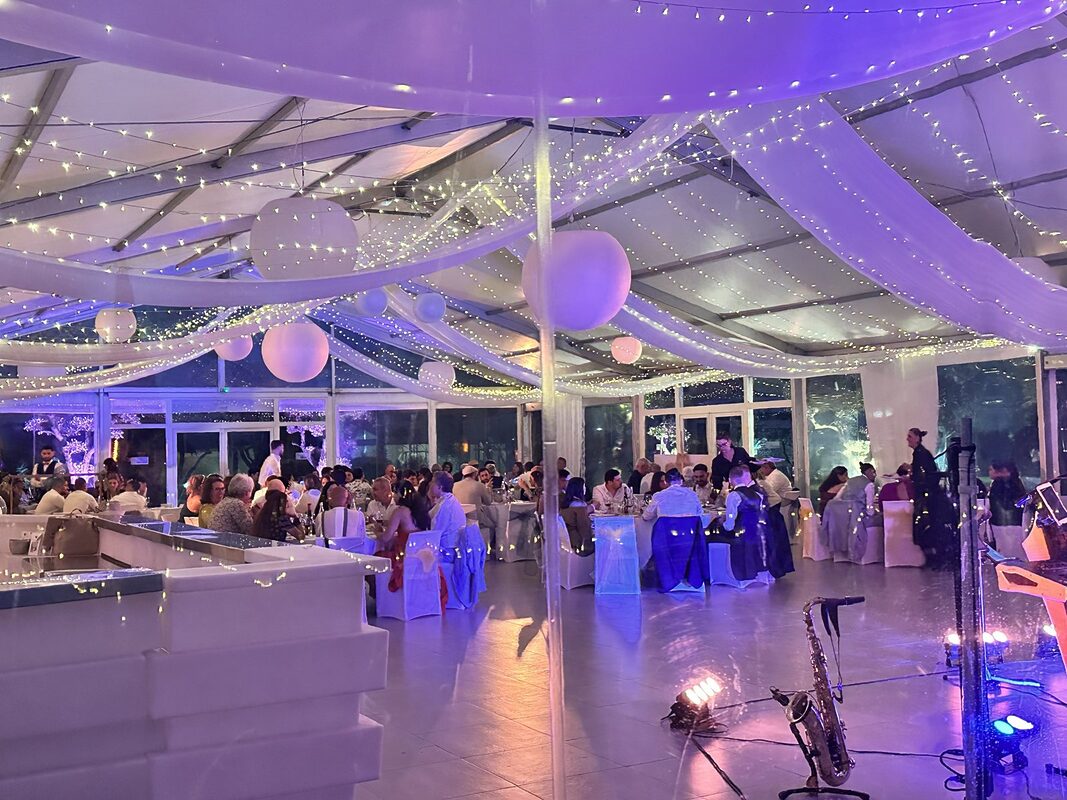 Dîner sous chapiteau, plafond drapé d’organza blanc et de guirlandes lumineuses, boules blanches suspendues, lumière violette