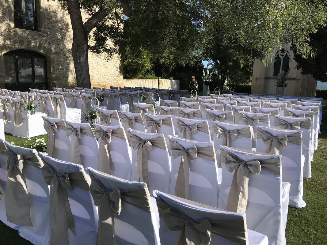 Allée de cérémonie entre des chaises housées de blanc à nœuds de satin taupe, crochets fleuris et colonnes de glaïeuls blancs, chapelle et étang en fond