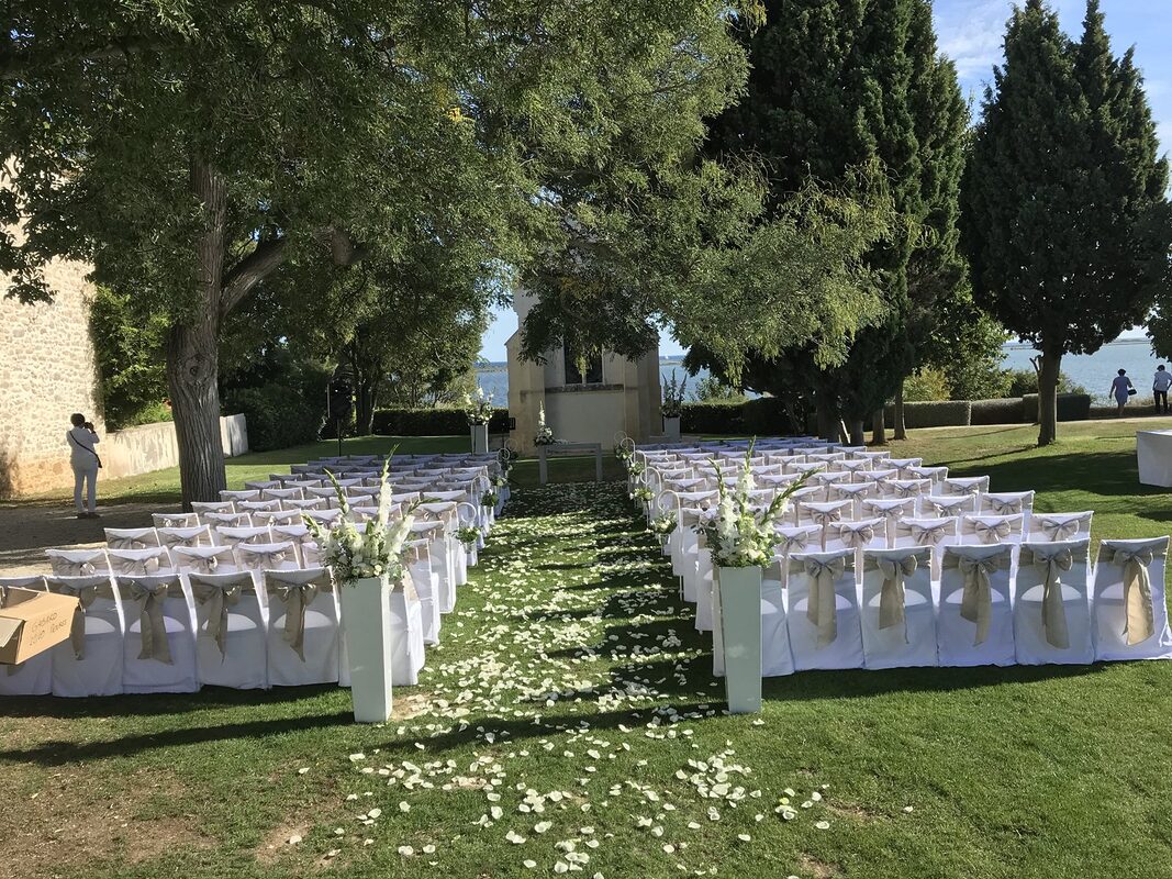 Cérémonie sous les arbres devant la chapelle du domaine : chaises housées à nœuds taupe, allée de pétales blancs bordée de hautes colonnes blanches fleuries