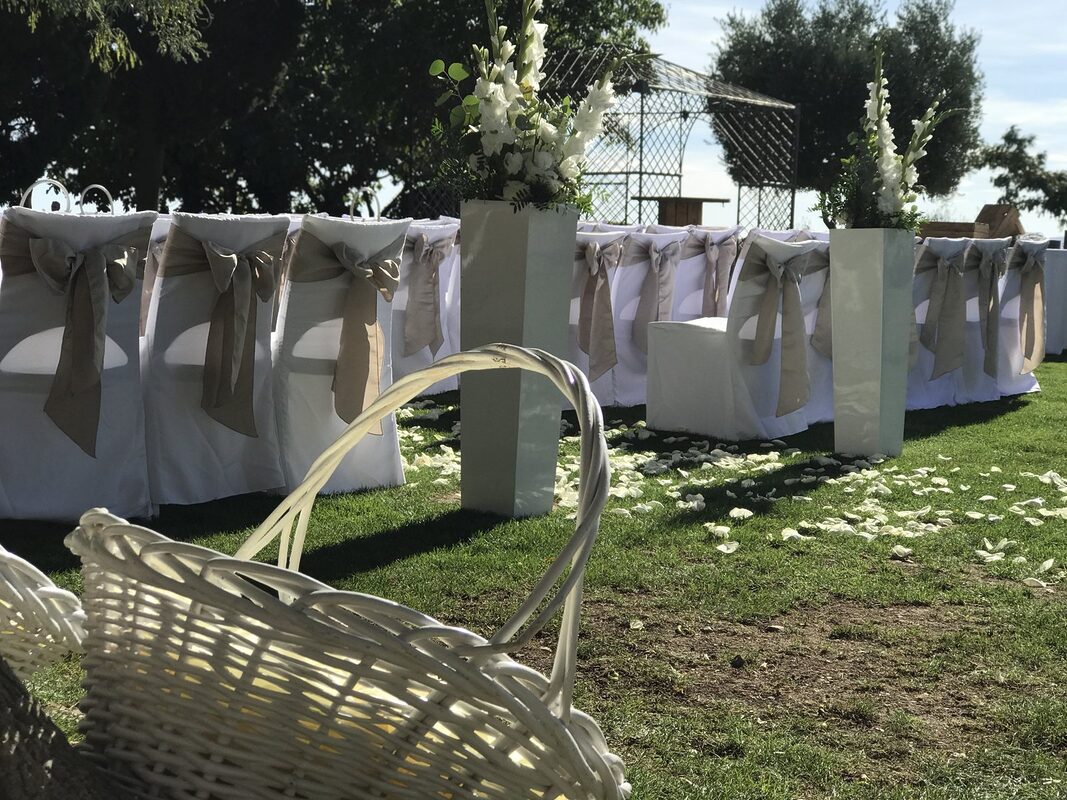 Panier en osier blanc pour les pétales au départ de l’allée, colonnes blanches fleuries et chaises à nœuds de satin
