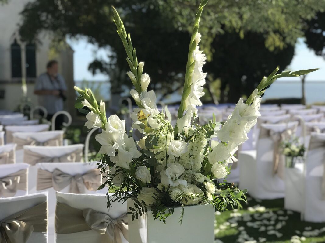Composition de glaïeuls, lisianthus et eucalyptus blancs sur colonne blanche, chaises à nœuds de satin taupe floues derrière