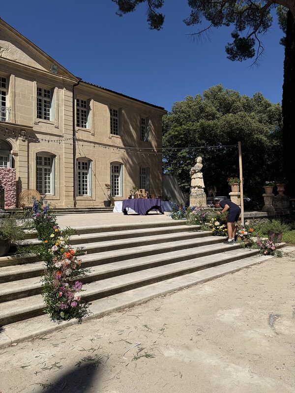 Grand escalier de pierre d’un château bordé de compositions florales, statues et guirlande lumineuse tendue sur la façade