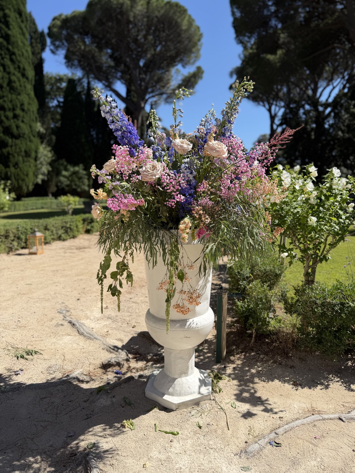 Vase Médicis blanc garni d’un bouquet champêtre rose et violet, posé au bord d’une allée de jardin bordée de cyprès