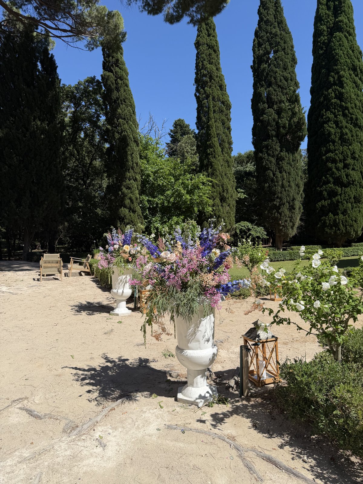 Vase Médicis blanc fleuri de delphiniums et de roses près d’un massif, lanternes au sol et cyprès dans le parc d’un château