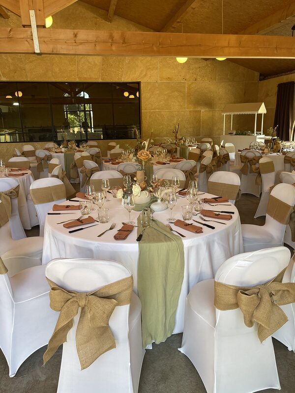 Tables rondes nappées de blanc, chemins de gaze vert sauge, serviettes terracotta et chaises housées à nœuds de jute