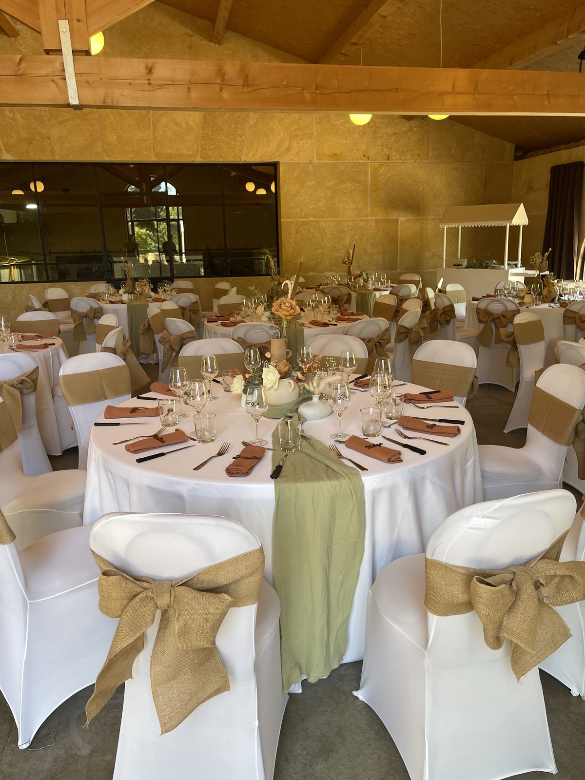 Tables rondes nappées de blanc, chemins de gaze vert sauge, serviettes terracotta et chaises housées à nœuds de jute