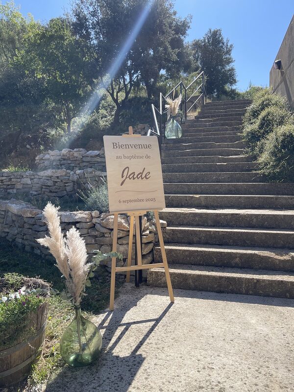 Panneau de bienvenue en bois sur chevalet au pied d’un escalier en pierre, dames-jeannes vertes garnies de pampas