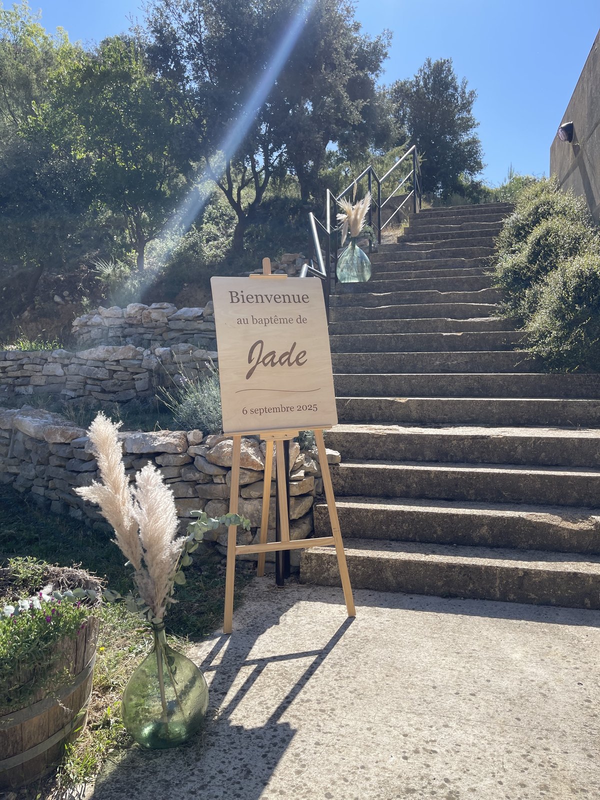 Panneau de bienvenue en bois sur chevalet au pied d’un escalier en pierre, dames-jeannes vertes garnies de pampas