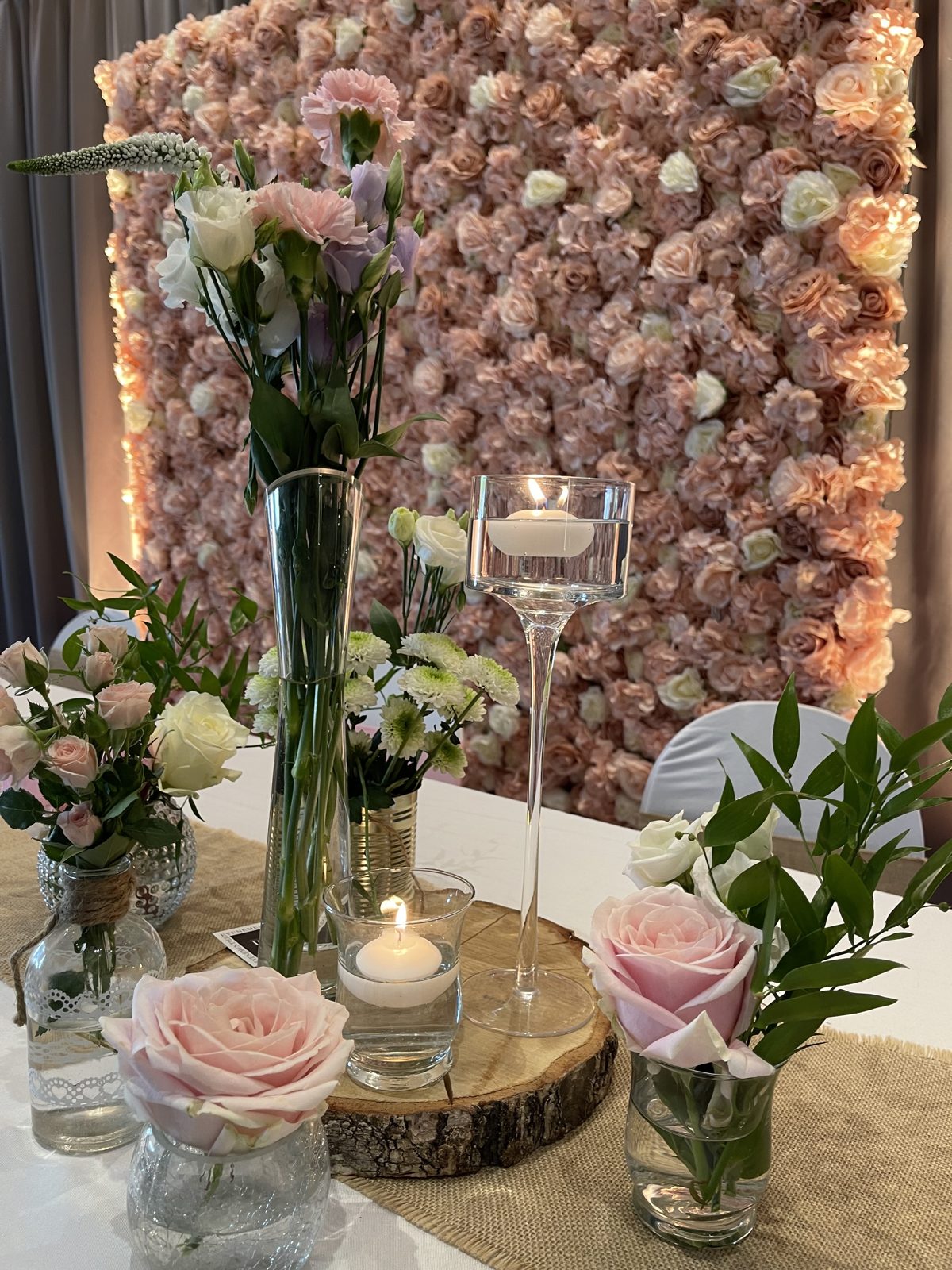 Centre de table champêtre : rondin de bois, roses pâles en vases de verre et bougies, mur floral rose en fond