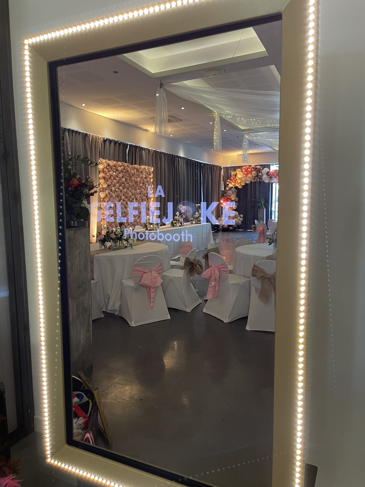 Borne photo miroir encadrée de LED reflétant une salle aux tables rondes, chaises à nœuds roses et arche de ballons