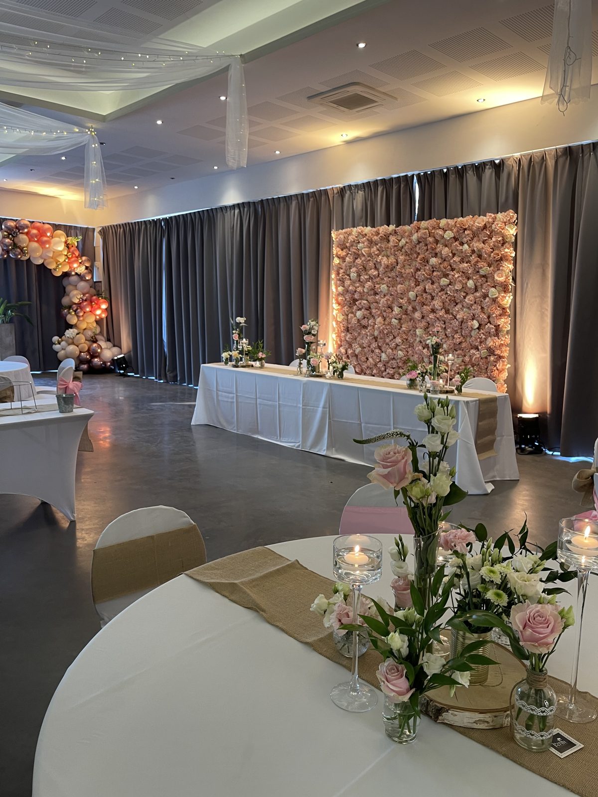 Salle de réception avec mur floral éclairé derrière la table d’honneur, table ronde fleurie et arche de ballons au fond