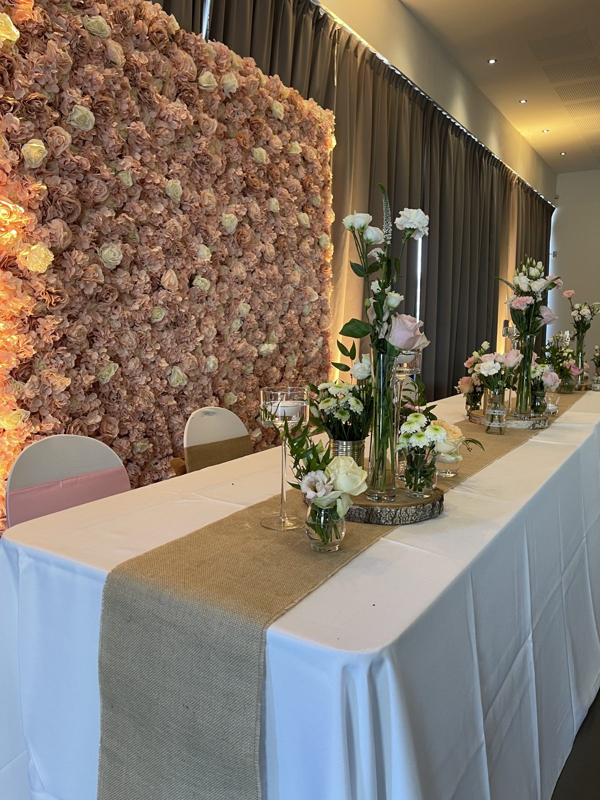 Table d’honneur nappée de blanc, chemin de table en jute et soliflores de roses devant un mur floral rose poudré