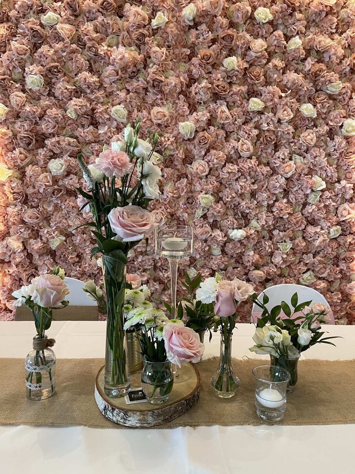 Mur floral rose et blanc en fond d’une table garnie de roses en vases de verre, rondin de bois et photophore