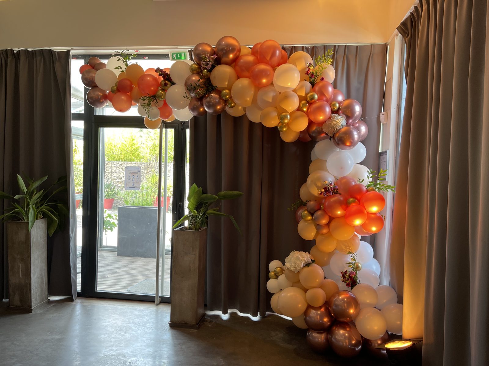 Arche de ballons organique blanc, pêche et cuivre fleurie encadrant une porte vitrée entre des rideaux gris