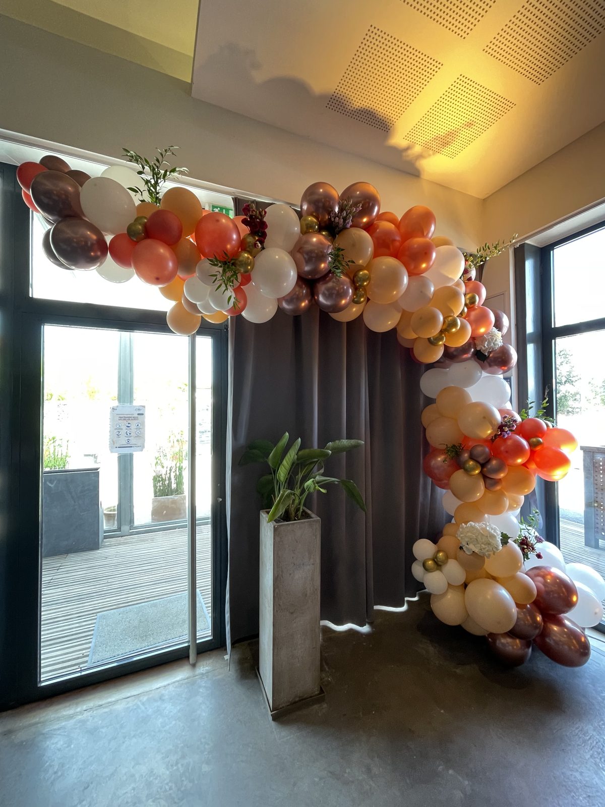 Arche de ballons blancs, pêche, cuivrés et dorés ornée de fleurs, installée devant un rideau gris près d’une baie vitrée