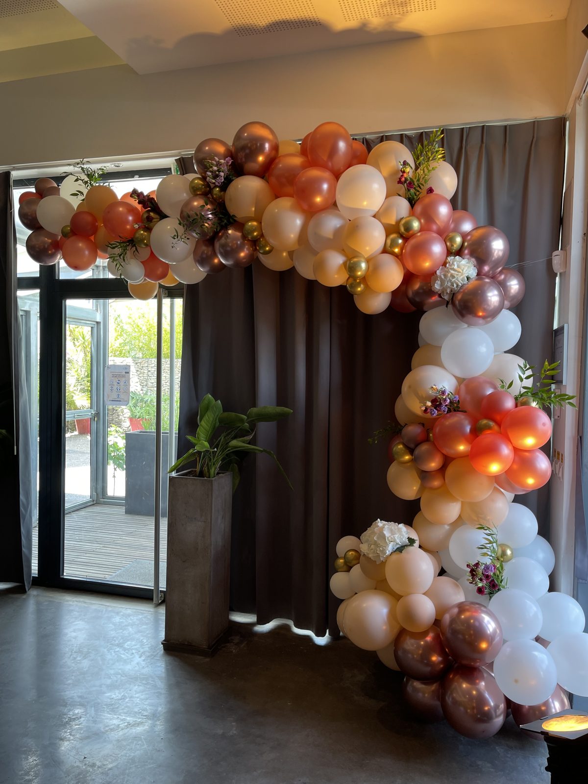 Détail d’une arche de ballons blancs, pêche, rose gold et dorés piquée de fleurs et de feuillage, salle en intérieur