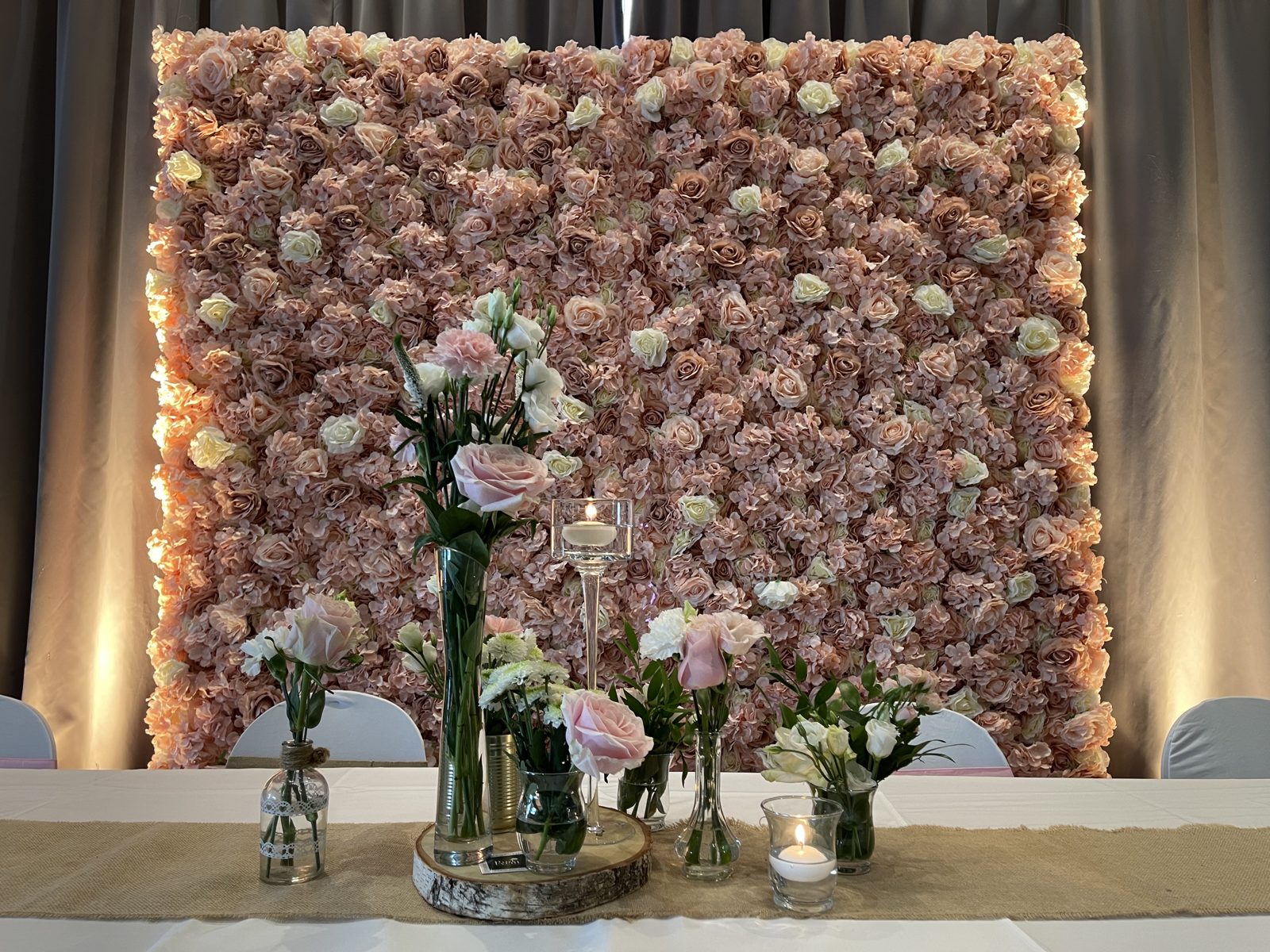 Gros plan de roses pâles en soliflores, bougie flottante sur haut pied et photophore devant un mur floral