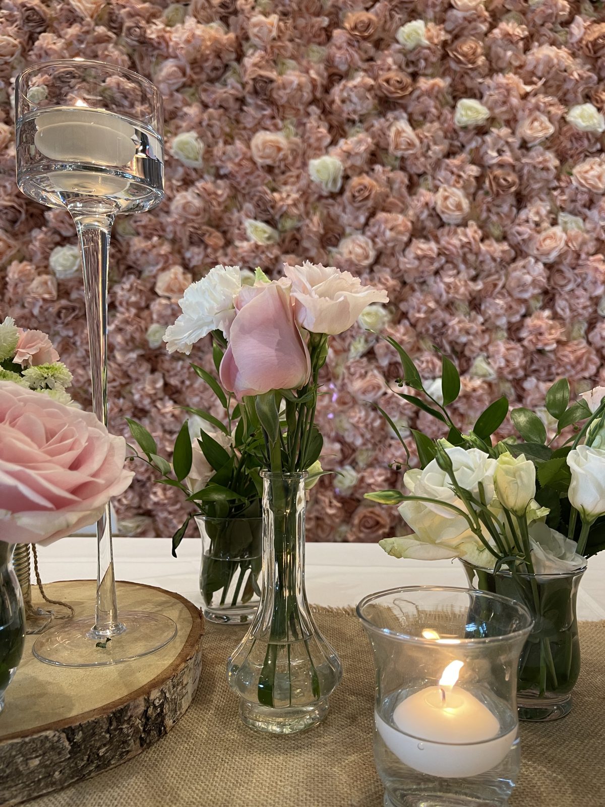 Mur floral rose poudré et blanc lumineux, table d’honneur avec chemin en jute, soliflores de roses et bougies